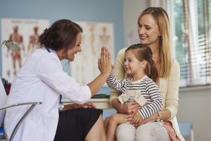 A doctor sitting in the office with two patients, a woman, and a child. The child and doctor are smiling while giving each other a high-five.